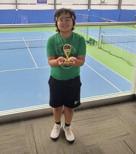 Aashil Image for Ed Brune Award_2 | Indianapolis Racquet Club Boy with glasses in a green shirt and black shorts, holding a yellow trophy inside a tennis facility with blue courts.