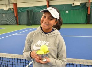 Izzy_May_2 | Indianapolis Racquet Club Smiling young woman in a gray hoodie and white visor holding a trophy filled with tennis balls on an indoor tennis court.