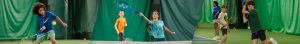 Group of children and teens playing tennis on an indoor court with green walls/curtains in the background, actively rallying. Use of multiple players suggests a practice or lesson scenario.